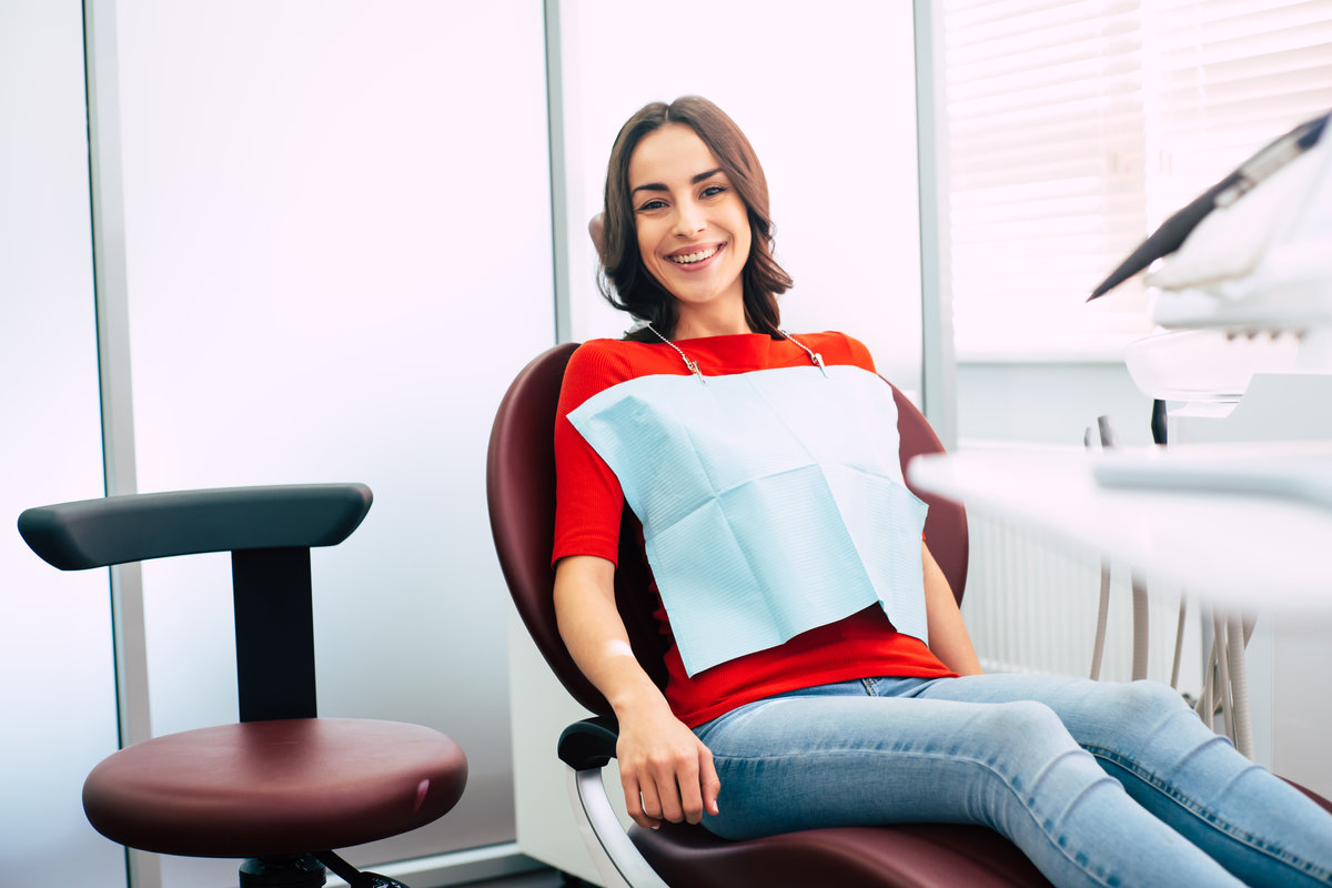 Ready to roll. Thin cute girl clothed in red sweater and denim jeans is sitting in the dental chair and waiting for the doctor with all of the pleasant emotions.