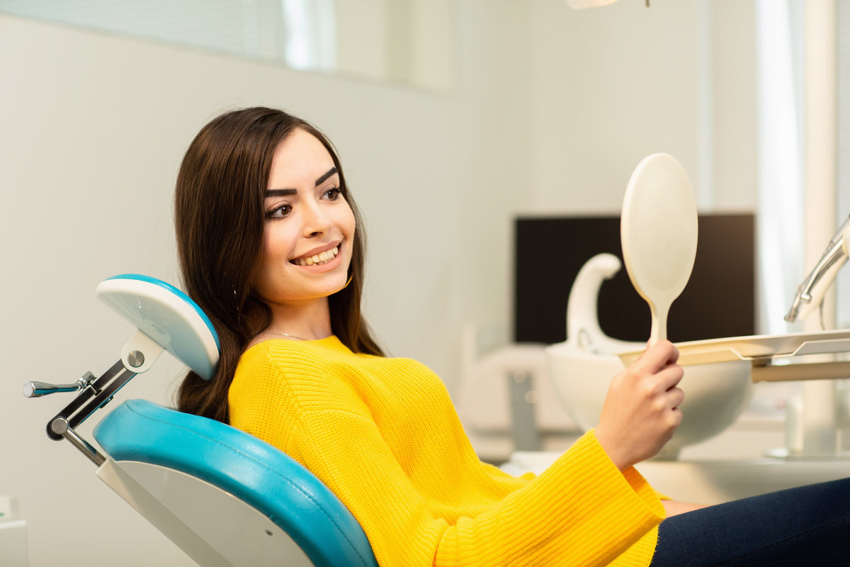 Young happy woman client looking at the mirror with toothy smile at the dental office.