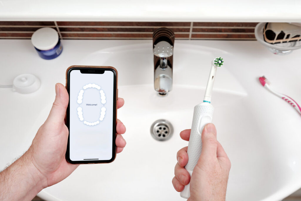 A man holds a smartphone and an electric toothbrush against bathroom sink. The smart electric toothbrush gives personalized dental care advice via a mobile app.